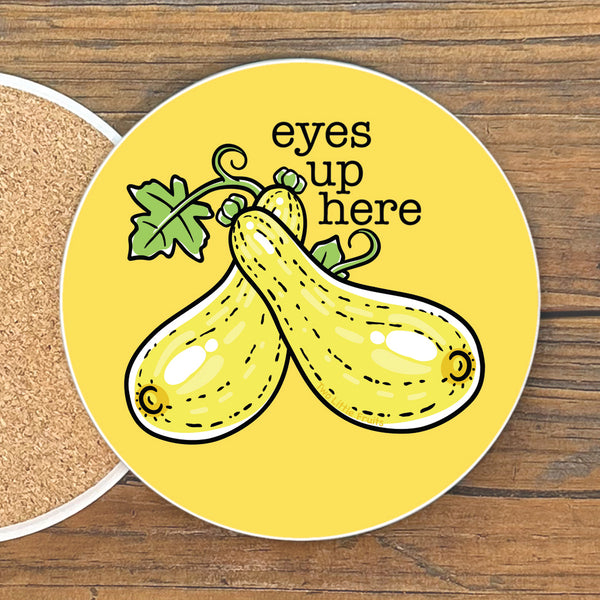 A yellow coaster with a cartoon illustration of two zucchini and the text "eyes up here" on it.