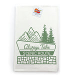 A white towel with a green graphic design depicting mountains, pine trees, and the text "Always Take the Scenic Route" printed on it.
