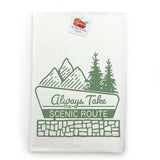A white towel with a green graphic design depicting mountains, pine trees, and the text "Always Take the Scenic Route" printed on it.