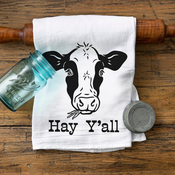 Hay Y’all cotton tea towel featuring a cow design, perfect for cow themed kitchen decor. The towel is placed on a wooden surface next to a glass jar and a round metal container, adding a rustic charm.