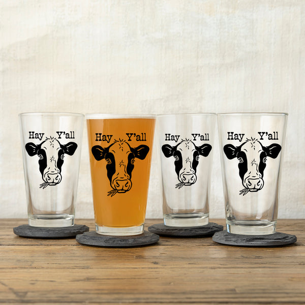 Variety of cow beer pint glasses labeled 'Hay Y'all', some filled with amber drinks, displayed on gray slate coasters for a stylish presentation.