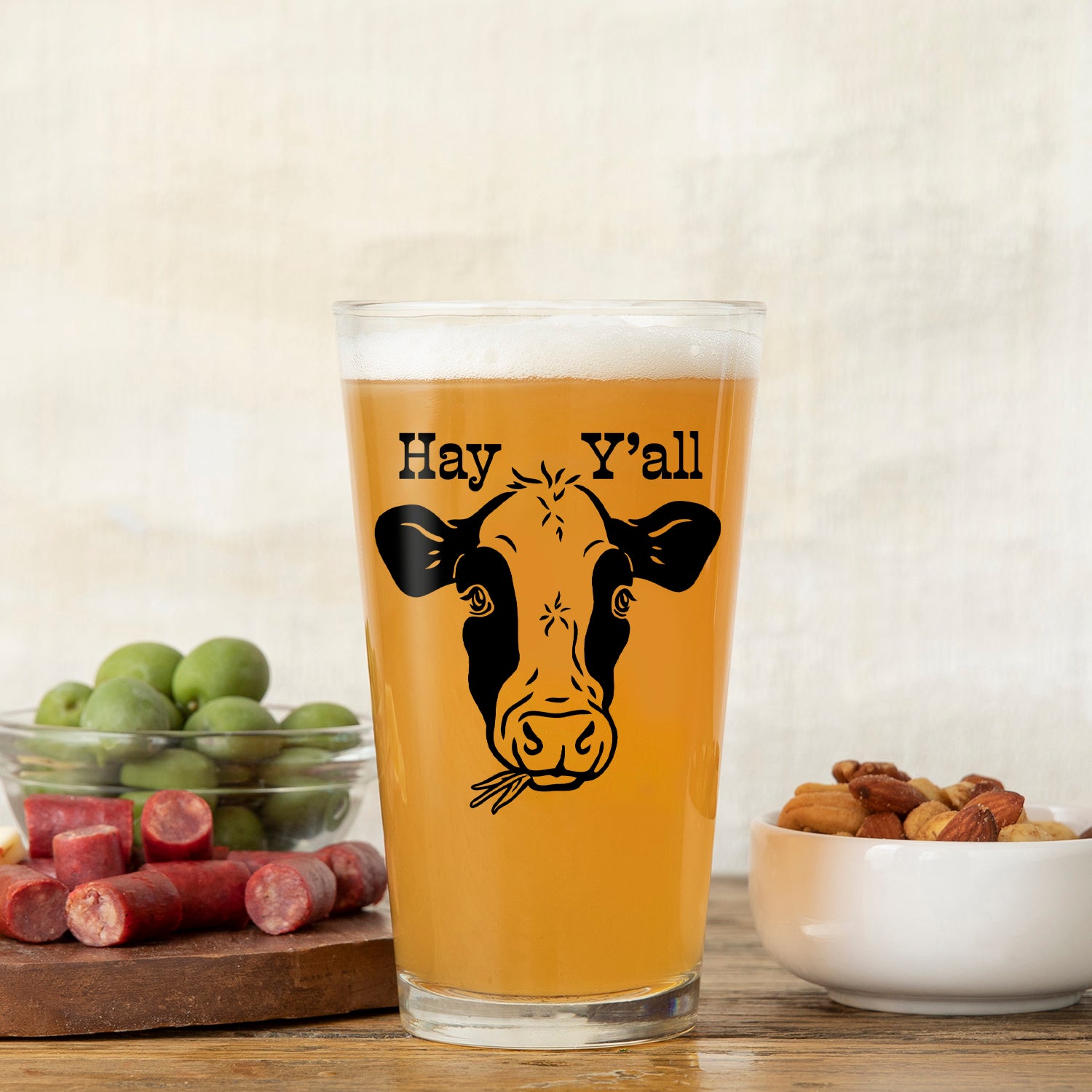 Close-up of a cow beer pint glass featuring the phrase 'Hay Y'all', showcasing its golden beverage alongside a plate of nuts and cured meats.