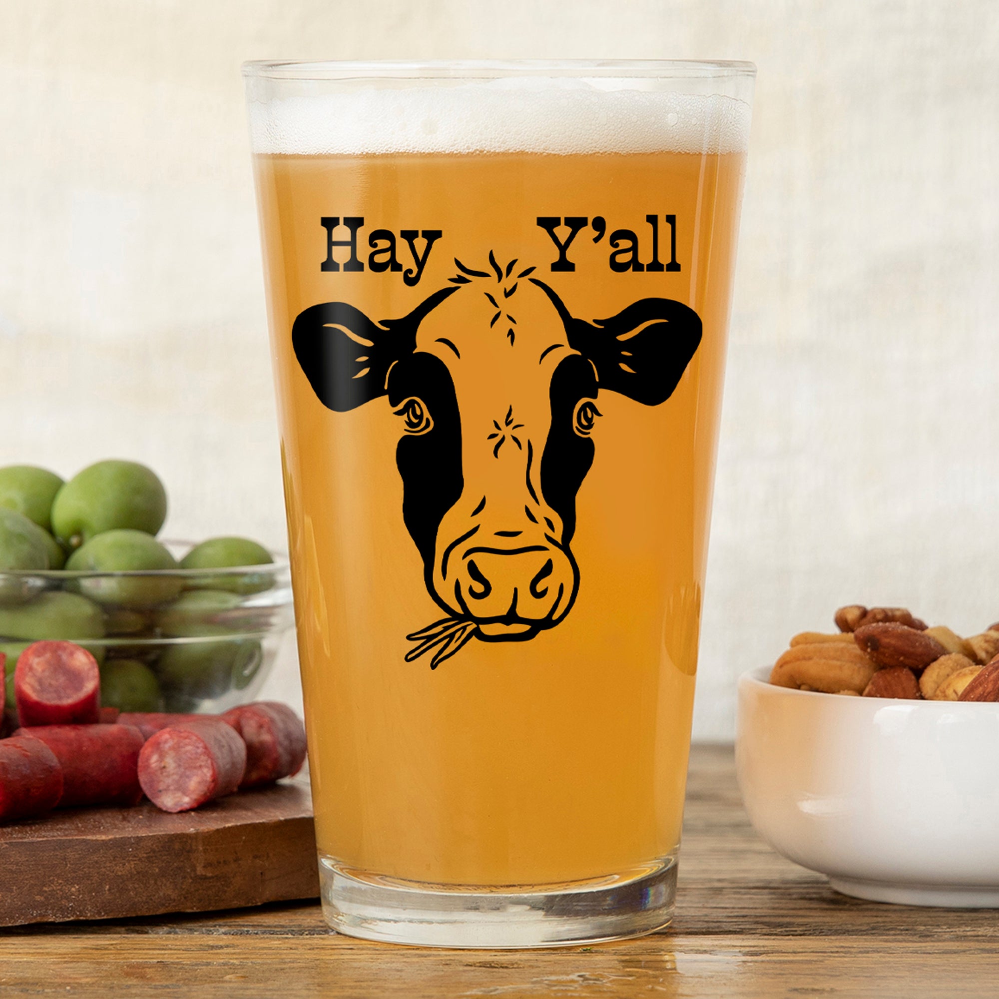 Cheerful cow beer pint glass with the words 'Hay Y'all' printed on it, filled with a golden drink, surrounded by green olives and snacks.