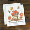 Mushroom dishcloth displayed on a rustic wooden surface, showcasing its vibrant red mushroom design along with flowers and a detailed label explaining its eco-friendly qualities.