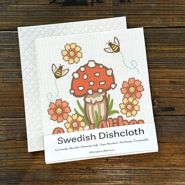 Mushroom dishcloth displayed on a rustic wooden surface, showcasing its vibrant red mushroom design along with flowers and a detailed label explaining its eco-friendly qualities.