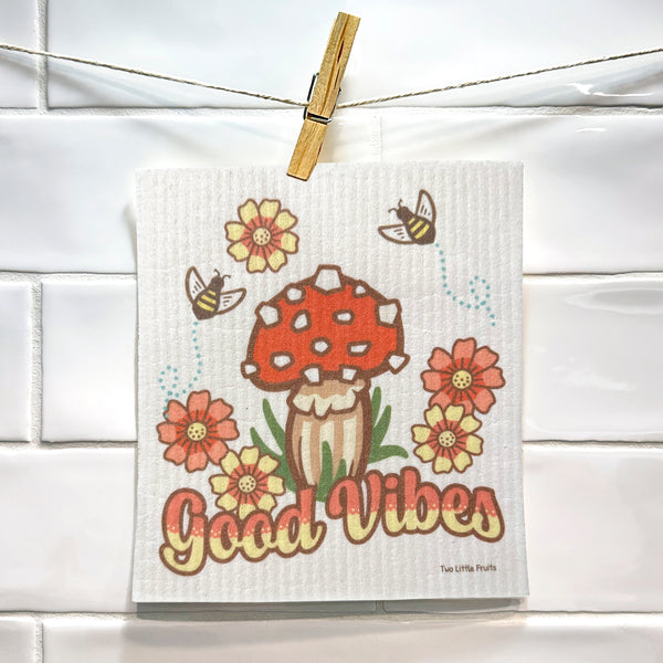 Mushroom dishcloth hung on a string with a clothespin, showcasing its playful design adorned with a red mushroom, flowers, and the caption 'Good Vibes' in a bright kitchen setting.