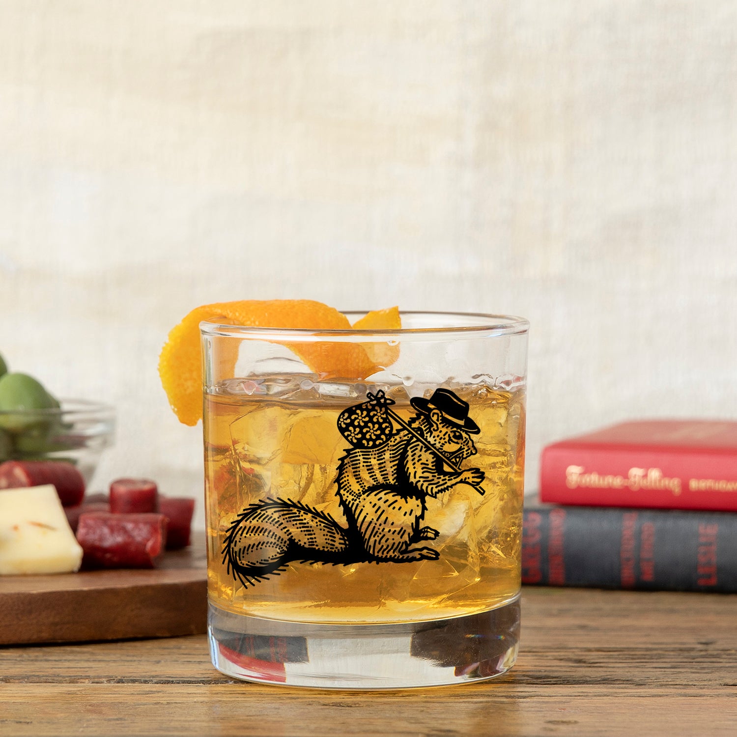 Another angle of a unique whiskey glass showcasing a hobo squirrel illustration, filled with a whiskey drink and orange garnish. Surrounded by cheese and a book on a wooden surface.