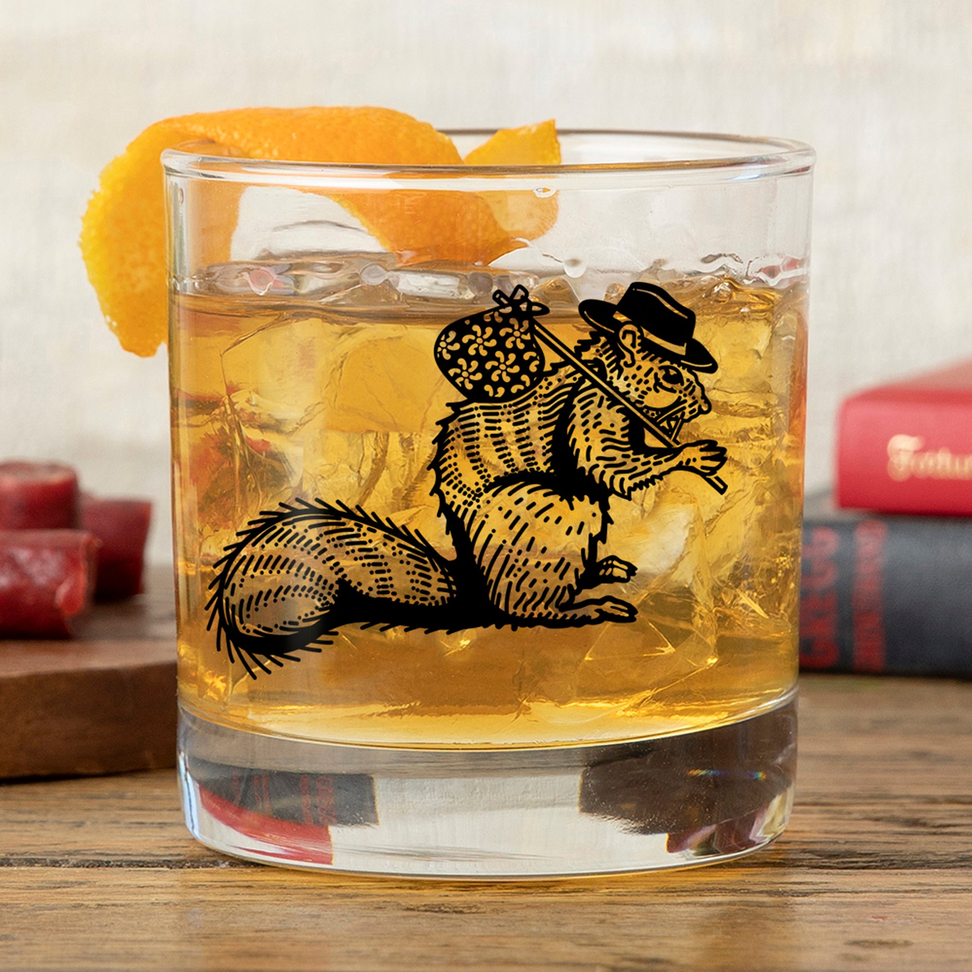 Unique whiskey glass filled with a golden drink, garnished with an orange peel. The glass features a whimsical hobo squirrel design, set against a rustic wooden table.