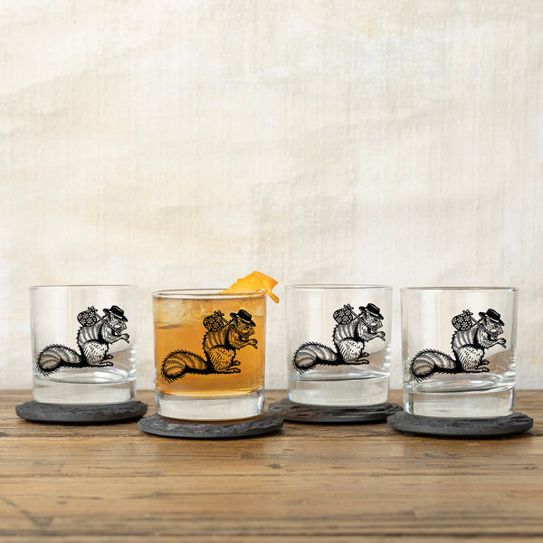 Four unique whiskey glasses lined up on slate coasters. Each glass displays a playful hobo squirrel design, with one filled with whiskey and garnished with orange.