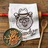 Howdy Bear Cowboy Kitchen Towel displayed on a wooden surface next to a rolling pin and a plate of baking chips, showcasing its fun design and practical use.
