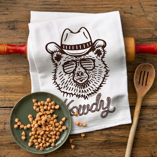 A cowboy bear kitchen towel featuring an artistic brown illustration of a bear wearing sunglasses and a cowboy hat, styled on a white cotton towel. The towel is placed on a rustic wooden surface surrounded by a green plate filled with small candy pieces and a wooden spoon.
