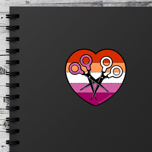 Lesbian Heart Scissors Sticker displayed on a black notebook, highlighting its bold and playful design. Perfect for personalizing your stationery, this sticker is a great representation of lesbian pride stickers.
