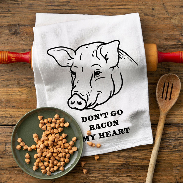 A humorous cotton tea towel featuring a playful pig design with the text 'DON'T GO BACON MY HEART.' The towel is displayed on a wooden surface alongside a rolling pin, a wooden spoon, and a plate of chocolate chips.