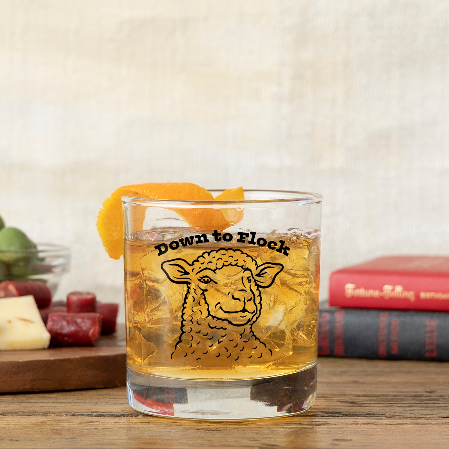 Funny Lamb Whiskey Glass filled with a drink, orange peel garnish, and surrounded by cheese and snack platters, showcasing a whimsical lamb design.