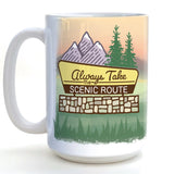 A white ceramic mug with a printed design depicting a scenic route, mountains, pine trees, and a sign that says "Always Take the Scenic Route".