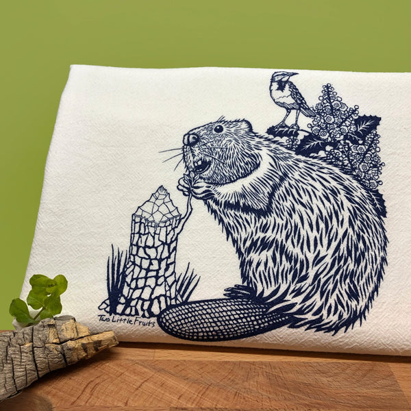 An artistic representation of a blue beaver with detailed flora on a beaver tea towel, set against a green background.