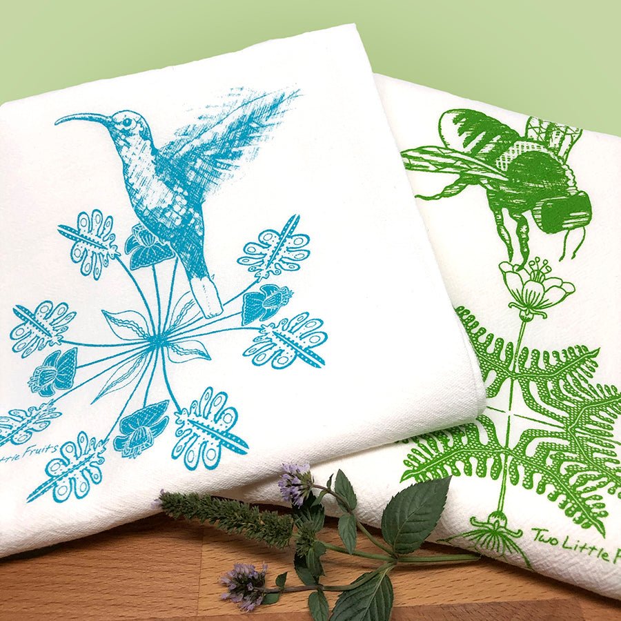 Colorful display of a turquoise hummingbird and a green bee tea towel, surrounded by herbs, on a kitchen countertop with a green backdrop.