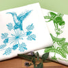Colorful display of a turquoise hummingbird and a green bee tea towel, surrounded by herbs, on a kitchen countertop with a green backdrop.