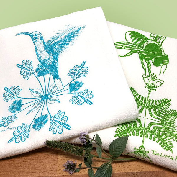Colorful display of a turquoise hummingbird and a green bee tea towel, surrounded by herbs, on a kitchen countertop with a green backdrop.