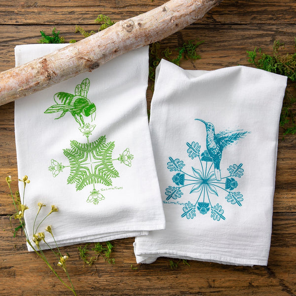 Cheerful kitchen towel set featuring a green bee illustration on the left and a turquoise hummingbird on the right, placed on a rustic wooden surface with sprigs of greenery.