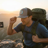 A man wearing a gray hat and backpack stands on a rocky mountain peak, with a backpack on his back and a backpack strap over his shoulder.
