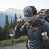 A man wearing a gray hat and backpack is walking on a trail in a mountainous area, with a backpack and water bottle attached to his backpack.