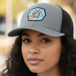 A young woman with long curly hair wearing a gray baseball cap with a graphic of a ram on it.