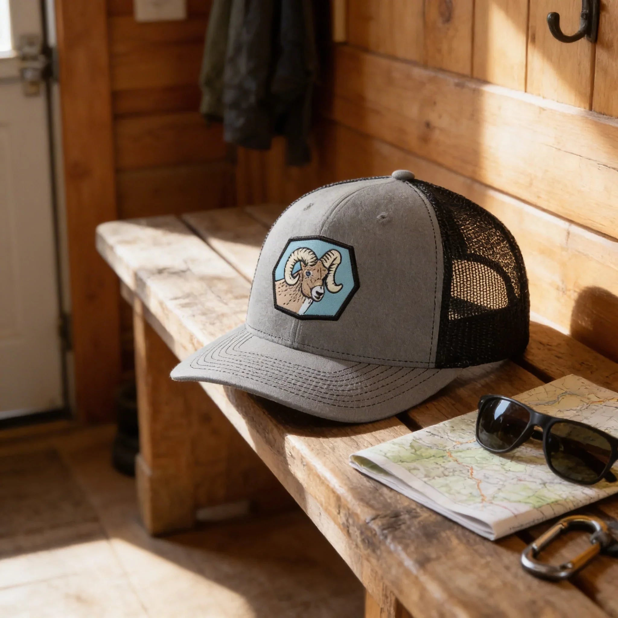 A gray baseball cap with a graphic of a ram sits on a wooden bench, accompanied by sunglasses, a map, and a backpack.