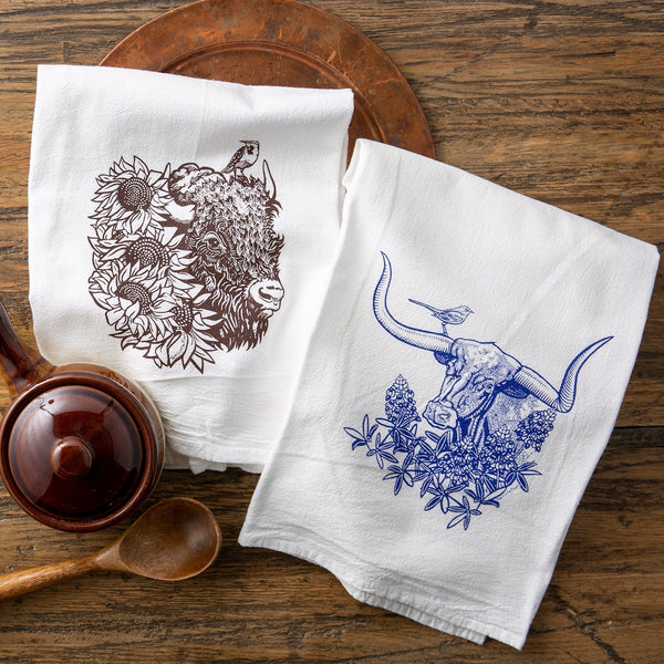 Cheerful colorful tea towel set featuring a brown bison design with sunflowers and a blue longhorn steer with bluebonnets on a wooden surface.