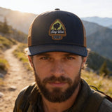 A man wearing a blue baseball cap with a patch on it, standing on a dirt path in a mountainous area.