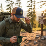 A man in a green jacket and a baseball cap is holding a green mug and adjusting his cap while standing on a dirt path surrounded by trees.