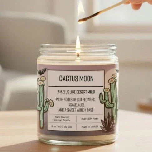 A hand is lighting a cactus-themed candle on a wooden table, with a potted plant visible in the background.