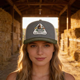 A woman wearing a gray baseball cap with a logo stands in front of a hay bale structure.