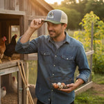 A man in a denim shirt and baseball cap stands in front of a wooden structure, holding a basket of eggs.