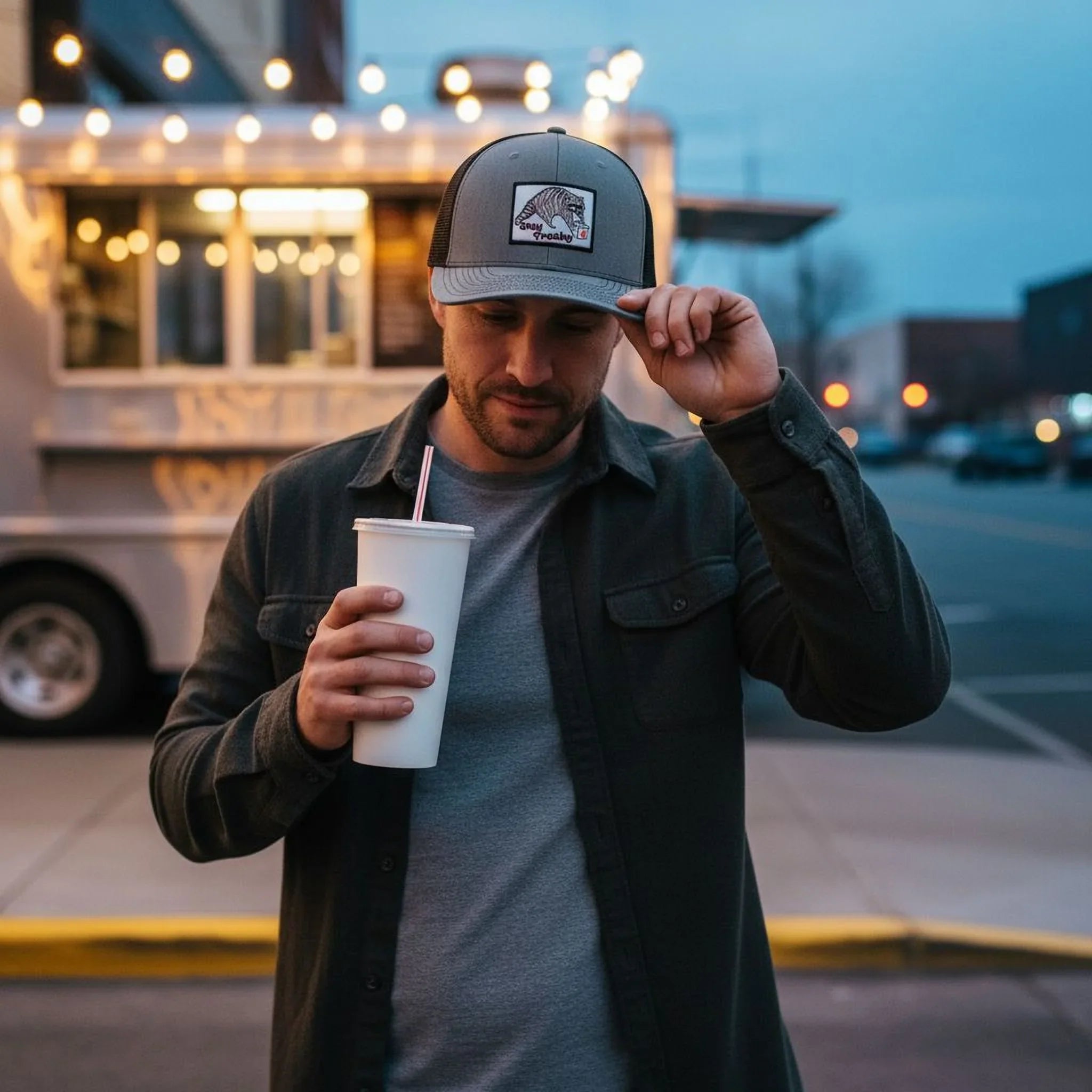 A man in a black jacket and gray cap stands on a city street, holding a white cup and looking down at it.