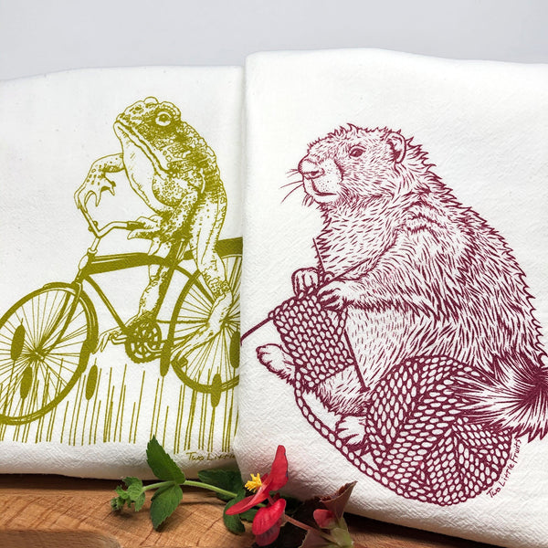 Close-up of Frog and Marmot Tea Towel Set showcasing a whimsical illustration of a moss green frog cycling and a raspberry knitting marmot, ideal for unique tea towel gifts.
