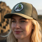 A woman wearing a green baseball cap with a cartoon frog riding a bicycle on it, standing in front of a rocky landscape.