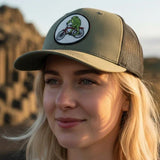 A woman wearing a green baseball cap with a cartoon frog riding a bicycle on it, standing in front of a rocky landscape.