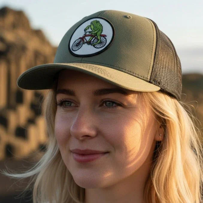 A woman wearing a green baseball cap with a cartoon frog riding a bicycle on it, standing in front of a rocky landscape.