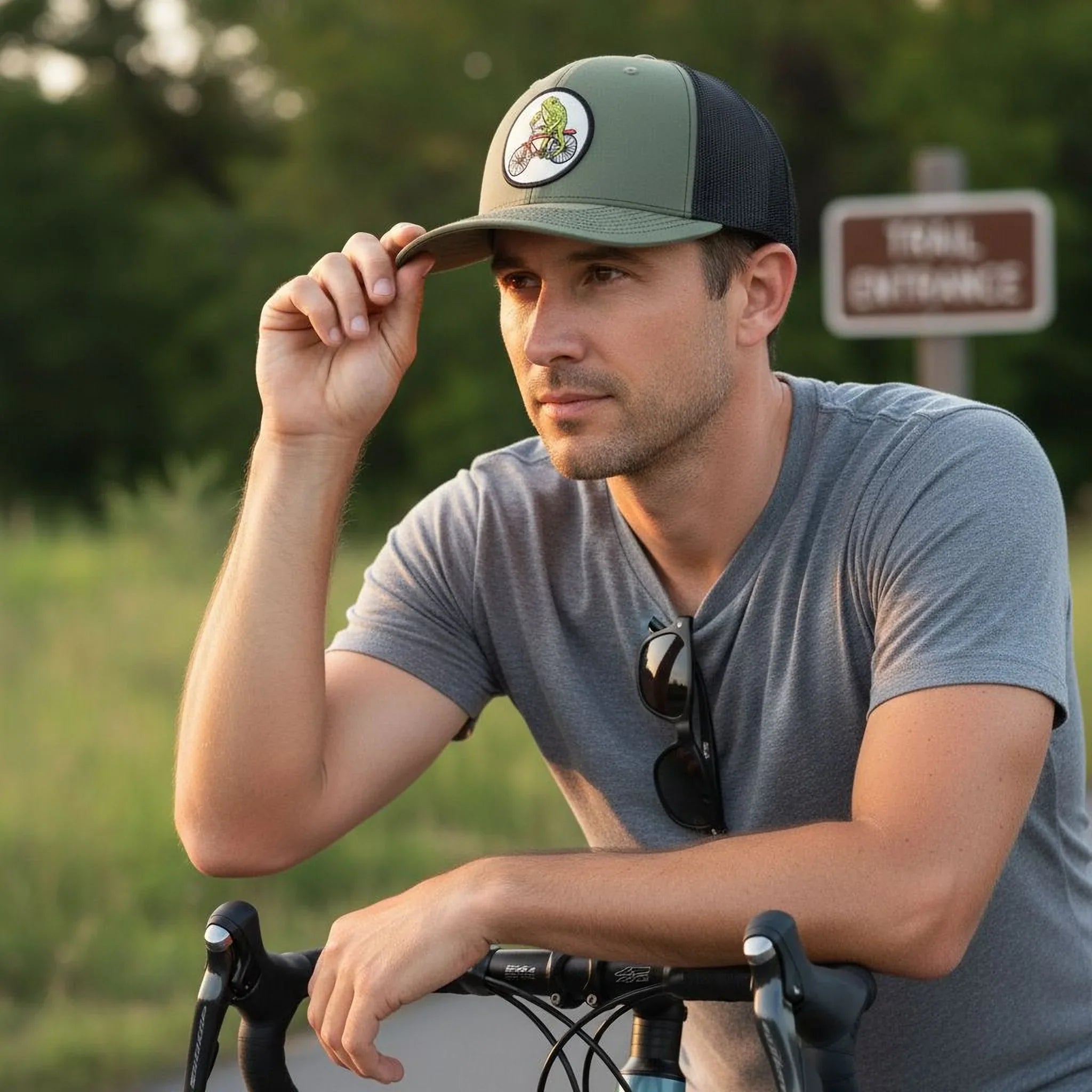 A man in a gray t-shirt and green baseball cap is leaning on a bicycle, with a trail sign visible in the background.