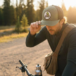 A man wearing a green hat with a patch on it is adjusting his hat while riding a bicycle on a dirt road surrounded by trees.