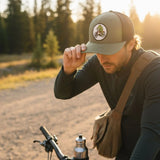 A man wearing a green hat with a patch on it is adjusting his hat while riding a bicycle on a dirt road surrounded by trees.