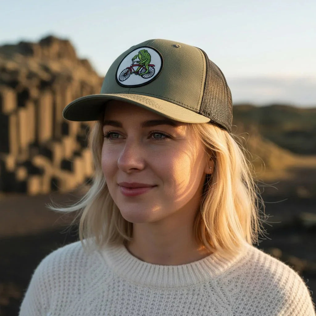 A woman wearing a green baseball cap with a cartoon frog riding a bicycle on it, standing in front of a rocky landscape.
