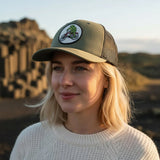 A woman wearing a green baseball cap with a cartoon frog riding a bicycle on it, standing in front of a rocky landscape.