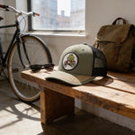 a wooden bench with a green baseball cap with a cartoon frog riding a bicycle on it, a pair of sunglasses, and a bicycle in the background.