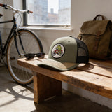 a wooden bench with a green baseball cap with a cartoon frog riding a bicycle on it, a pair of sunglasses, and a bicycle in the background.