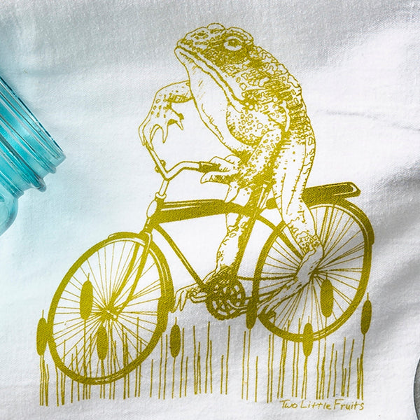 Adorable Frog Tea Towel featuring a moss green frog on a vintage cruiser bicycle, made from 100% cotton, eco-friendly and perfect for kitchen decor.