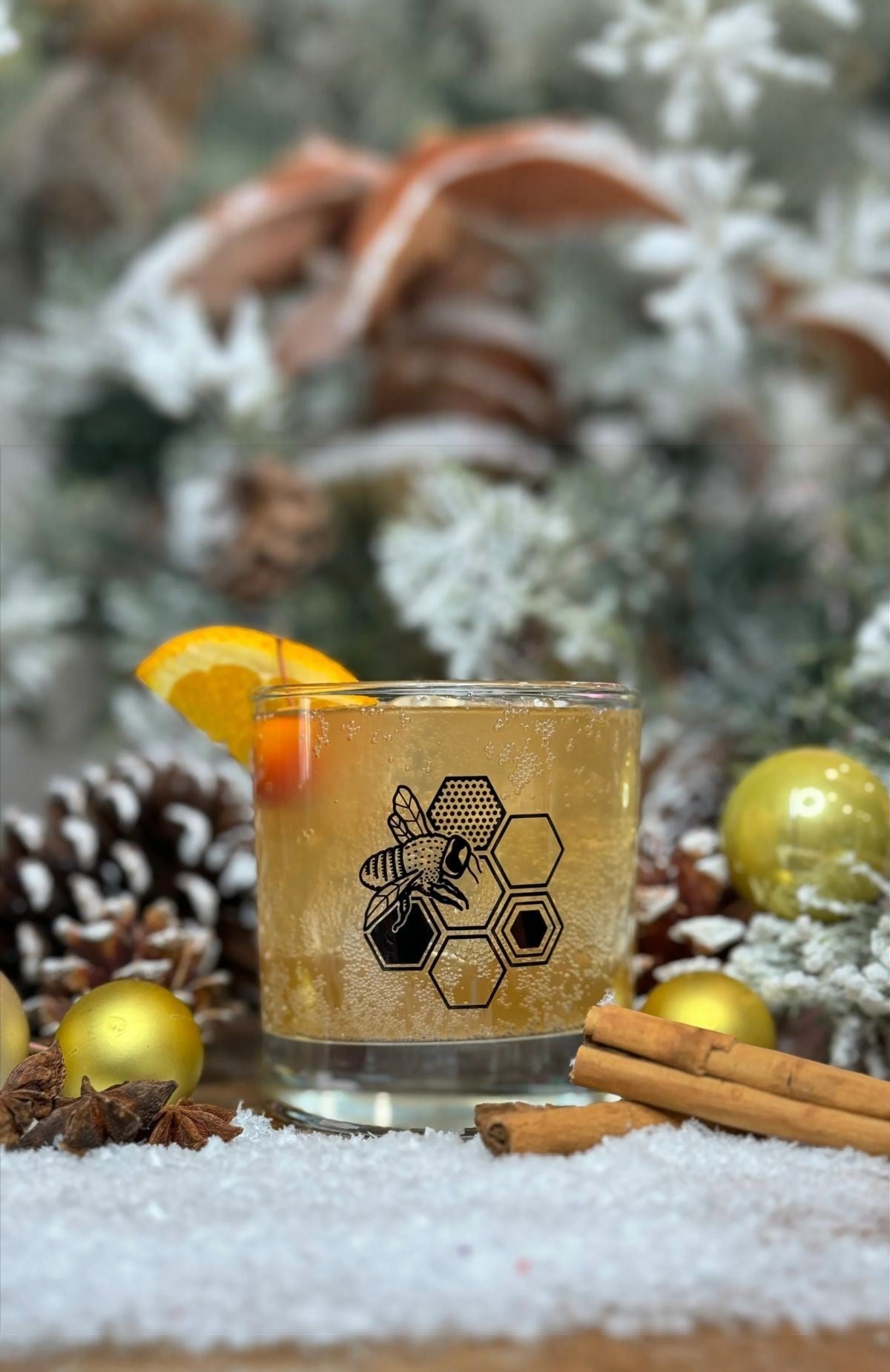 Honeybee rocks glass with an orange slice, pinecones and cinnamon in snow
