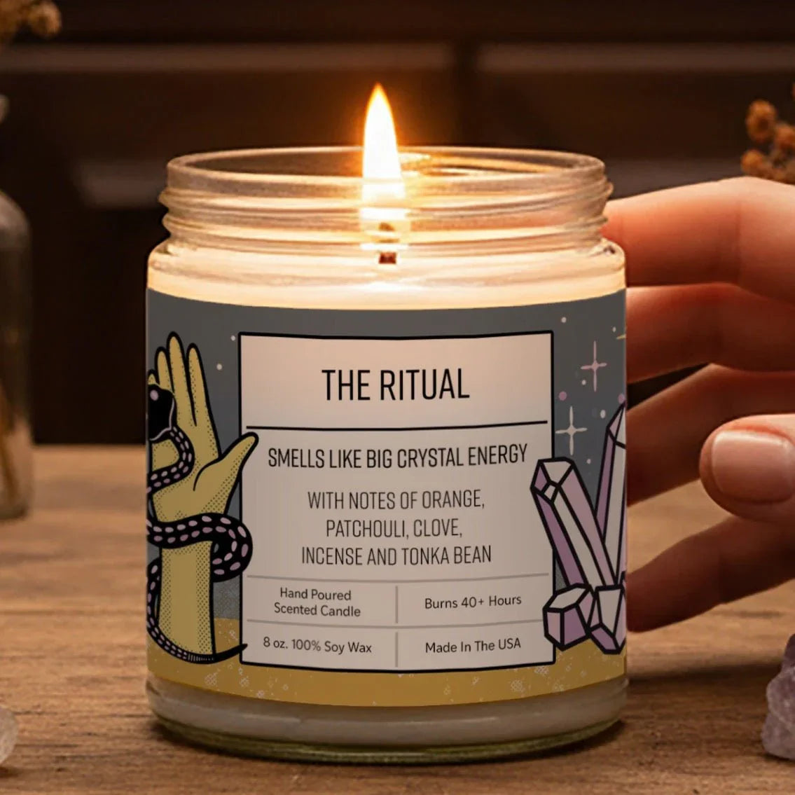 A hand is holding a lit candle labeled "THE RITUAL" on a wooden surface, surrounded by various crystals, dried flowers, and a bird statue.