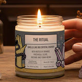 A hand is holding a lit candle labeled "THE RITUAL" on a wooden surface, surrounded by various crystals, dried flowers, and a bird statue.
